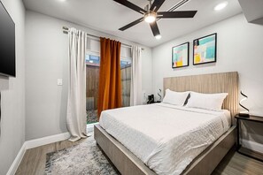 Ein modernes Schlafzimmer mit Bett, Deckenventilator, Kunstwerken und Blick durch eine Glastür.
