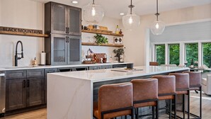 Una cocina moderna con una isla grande, taburetes de bar, luces colgantes y vistas a la vegetación.