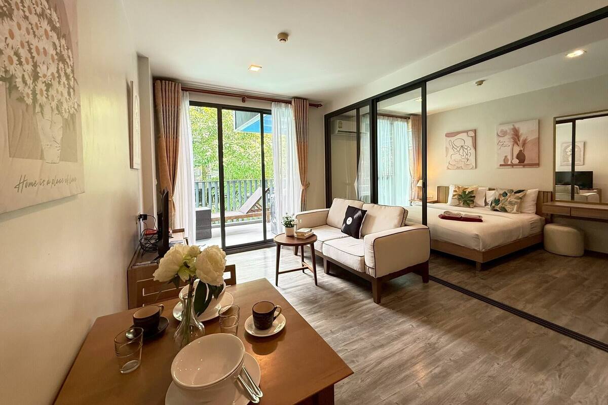 A spacious living room with a sofa, a dining table with a vase of flowers, and a large sliding glass door leading to a balcony.