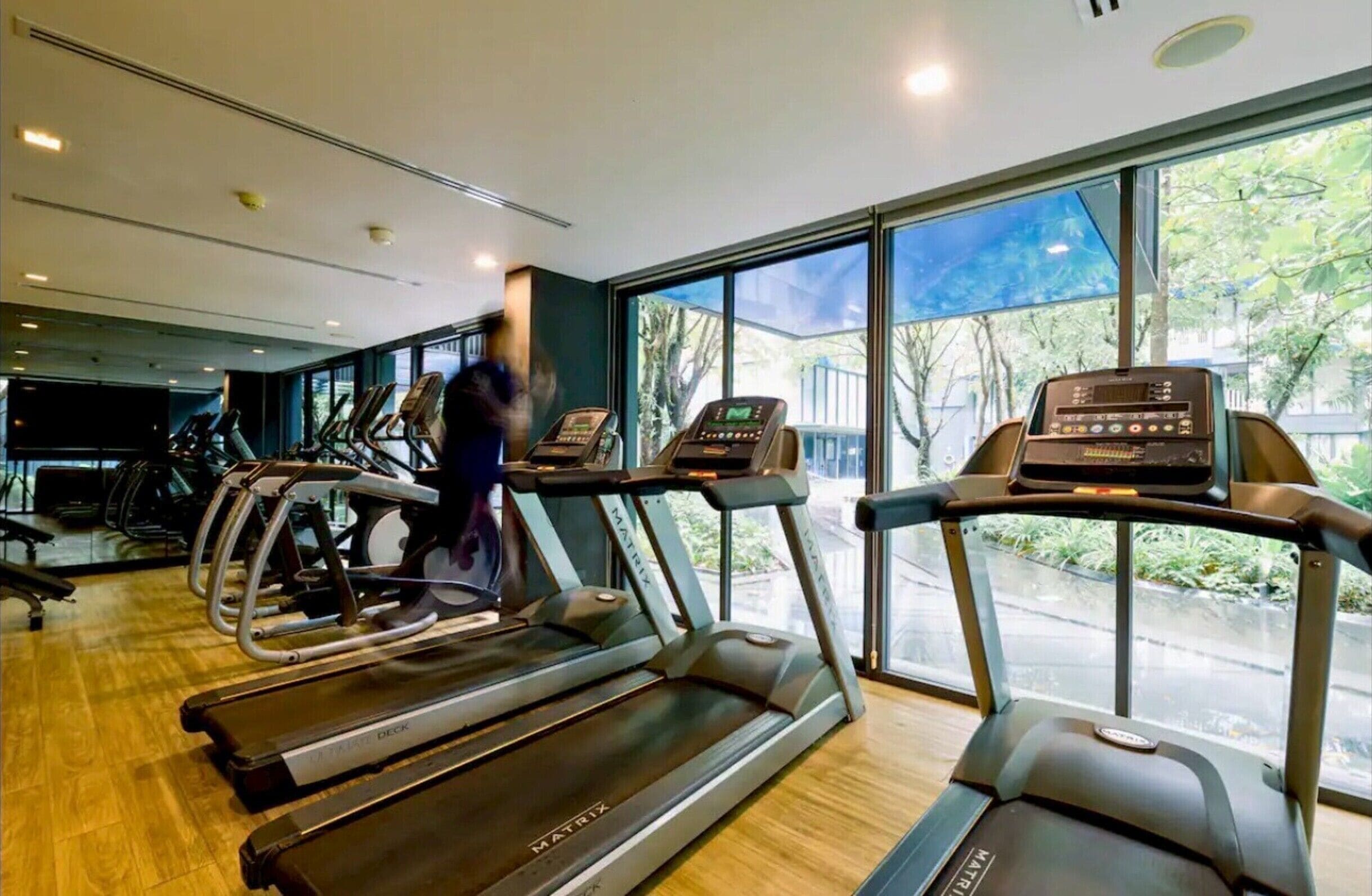 A gym with treadmills and exercise bikes, large windows with a view of trees, and a person walking on the treadmill.