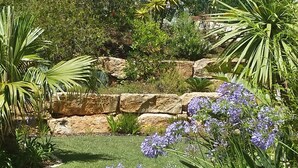 A well-maintained garden with a variety of plants and flowers, a stone retaining wall, and a clear blue sky. - Spacious Studio at Estrela Da Luz, pools, indoor spa and gym. (Luz)