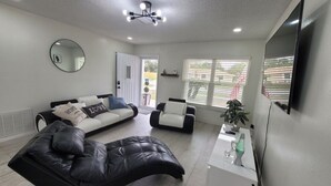 A modern living room with a black leather sofa, a white sofa, and a black leather couch. There is a flat-screen TV mounted on the wall, a ceiling light fixture, and a window with blinds. - Tranquil 2-bedroom house with AC in Spring Hill (Spring Hill)