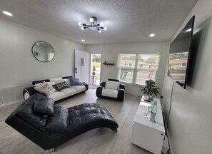 Ein modernes Wohnzimmer mit einem schwarzen Ledersofa, einem weißen Sofa und einer schwarzen Lederliege. An der Wand ist ein Flachbildfernseher angebracht, eine Deckenleuchte sorgt für Licht und ein Fenster mit Jalousien.