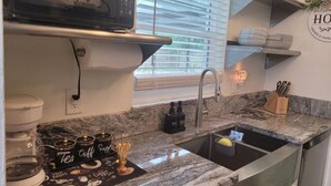 A kitchen with a granite countertop, a coffee maker, and a sink. There are shelves above the counter holding plates and glasses. - Tranquil 2-bedroom house with AC in Spring Hill (Spring Hill)