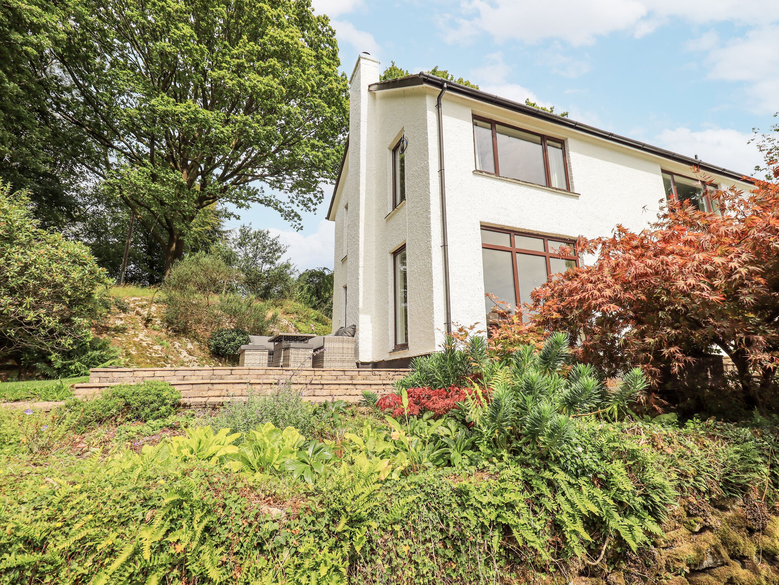 Une maison à deux étages, à l’extérieur blanc, avec un grand arbre sur la gauche et un jardin où l’on peut voir différentes plantes au premier plan.