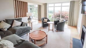 Een moderne woonkamer met een grijze bank, twee fauteuils, een houten salontafel en een lamp. Grote ramen bieden uitzicht op bomen en een landschap in de verte.