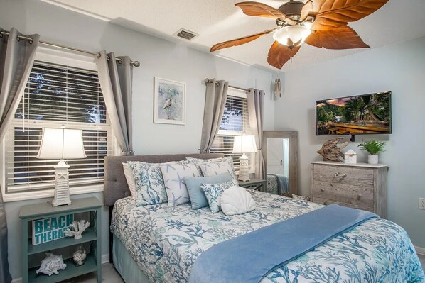 A bedroom with a bed, two bedside tables with lamps, a ceiling fan, a dresser, and a wall-mounted TV.