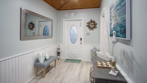 A hallway with a wooden ceiling, a door with a frosted glass panel, a bench, a small table with a tray of cookies, and a painting on the wall.