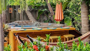 A wooden hot tub with a table and chairs, surrounded by lush greenery and a hammock.