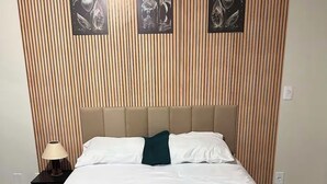 Una cama con sábanas blancas y una manta verde azulada, una mesita de noche con una lámpara, y una pared con listones de madera verticales y dos cuadros con motivos florales.