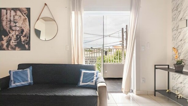 Una sala de estar moderna con un sofá negro, dos almohadas azules, un espejo redondo y un cuadro enmarcado con leones. A la derecha, una pequeña mesa con una planta.