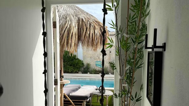 Une pièce avec un fauteuil suspendu, une grande fenêtre, une plante en bambou et une vue sur une piscine et du mobilier d’extérieur.