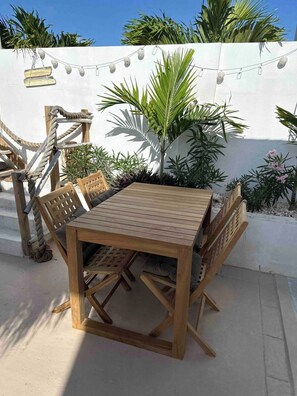 A wooden outdoor table with chairs, surrounded by plants and a white wall.