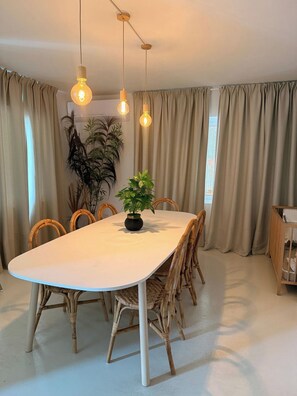 Een eethoek met een witte tafel, rieten stoelen, een potplant en hangende pendantlampen.