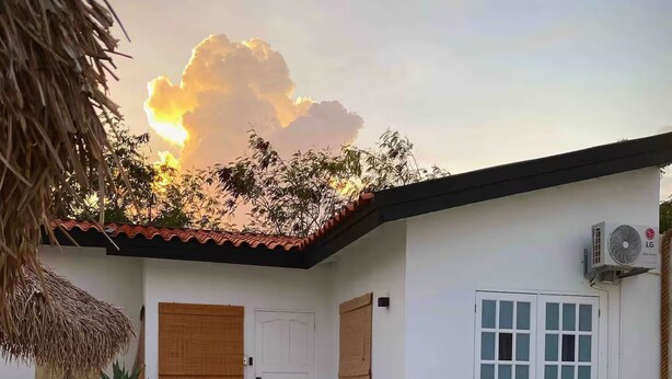 A poolside area with a thatched roof, a white building with a tiled roof, and a clear sky. - Palm Beach VacaySuites - Garden Suite - Poolside (Palm Beach)