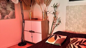 Un dormitorio con una colcha de cama estampada en tonos oscuros, un aparador rosa, una lámpara de pie y plantas decorativas.