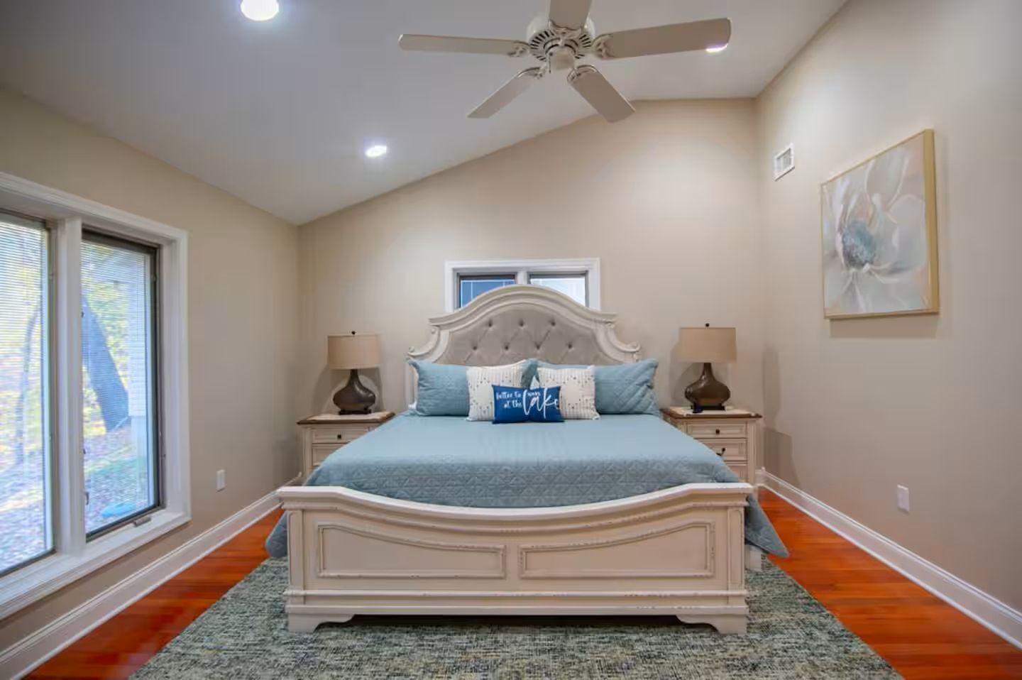 A bedroom with a large bed, two bedside tables, a ceiling fan, a window, and a painting on the wall.