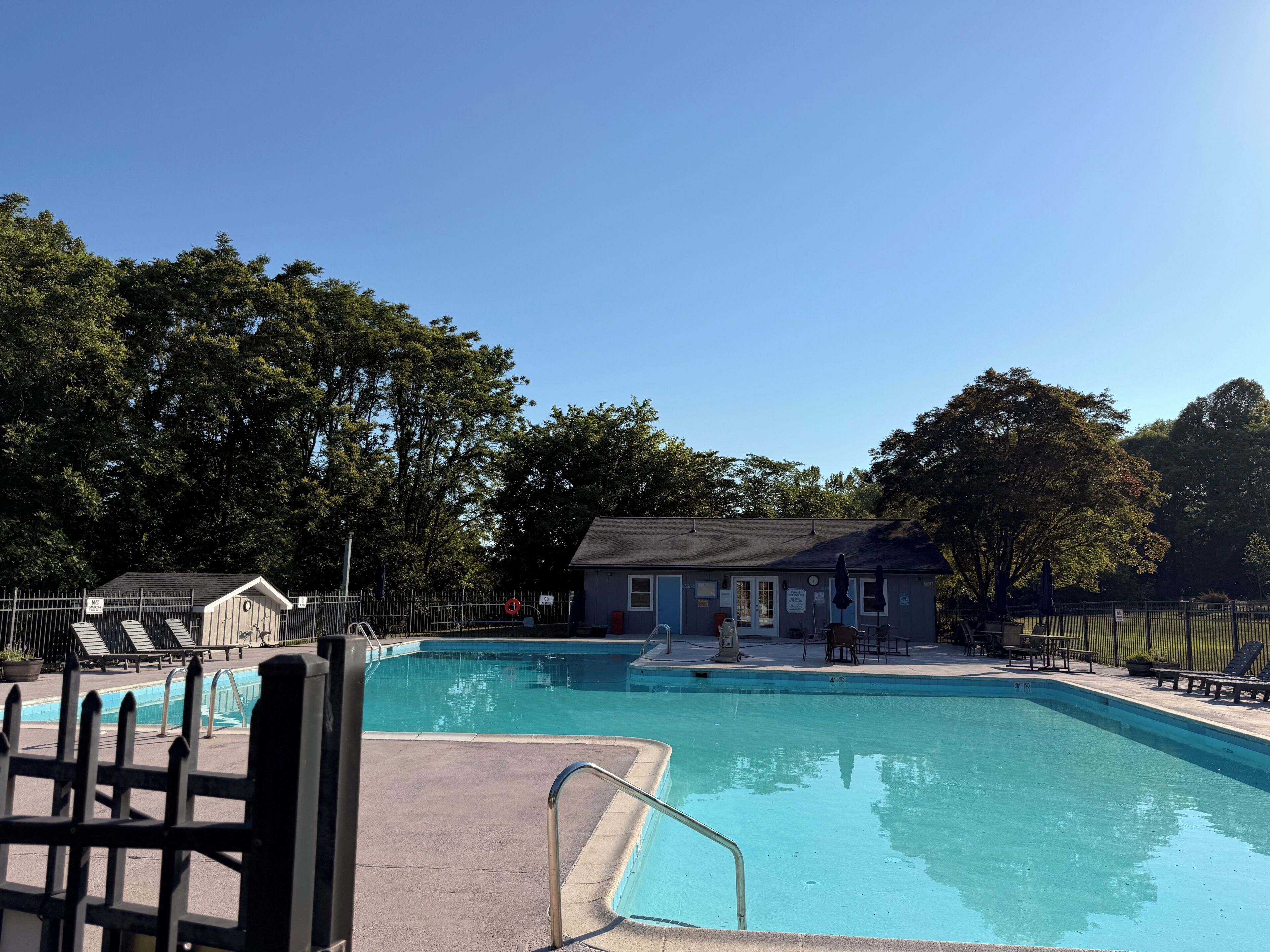 A swimming pool with a fenced deck, lounge chairs, and a small building in the background.
