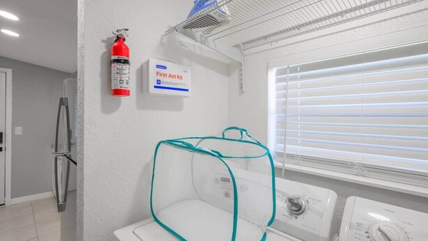 Een wasserette met een wasmachine, een brandblusser, een verbanddoos en een raam met zonwering.