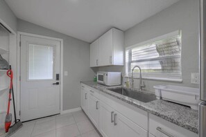 Une cuisine avec des meubles blancs, un plan de travail en granit, un four à micro-ondes, un évier et une fenêtre avec des stores.