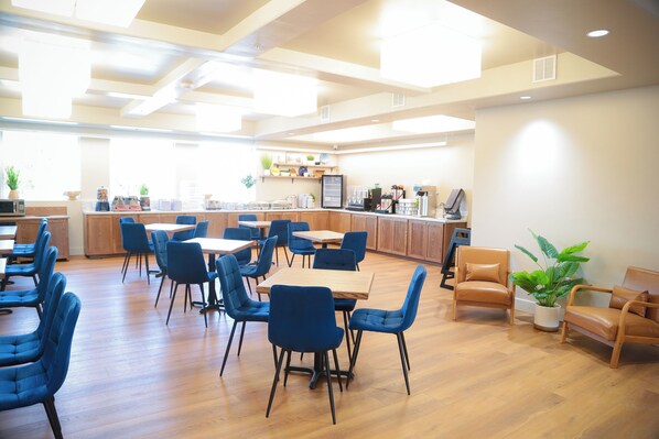 A spacious dining area with wooden tables and blue chairs, a kitchen area with appliances, and a potted plant. - Splash Canyon Hotel (Twin Falls)