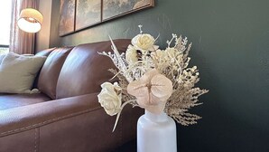 A living room with a brown leather sofa, a white vase with flowers, and a painting on the wall.