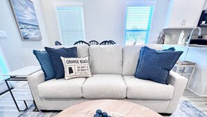 Un salon avec un canapé d’angle beige, une table basse ronde ornée d’un centre de table bleu et blanc, et un tableau représentant une plage accroché au mur.