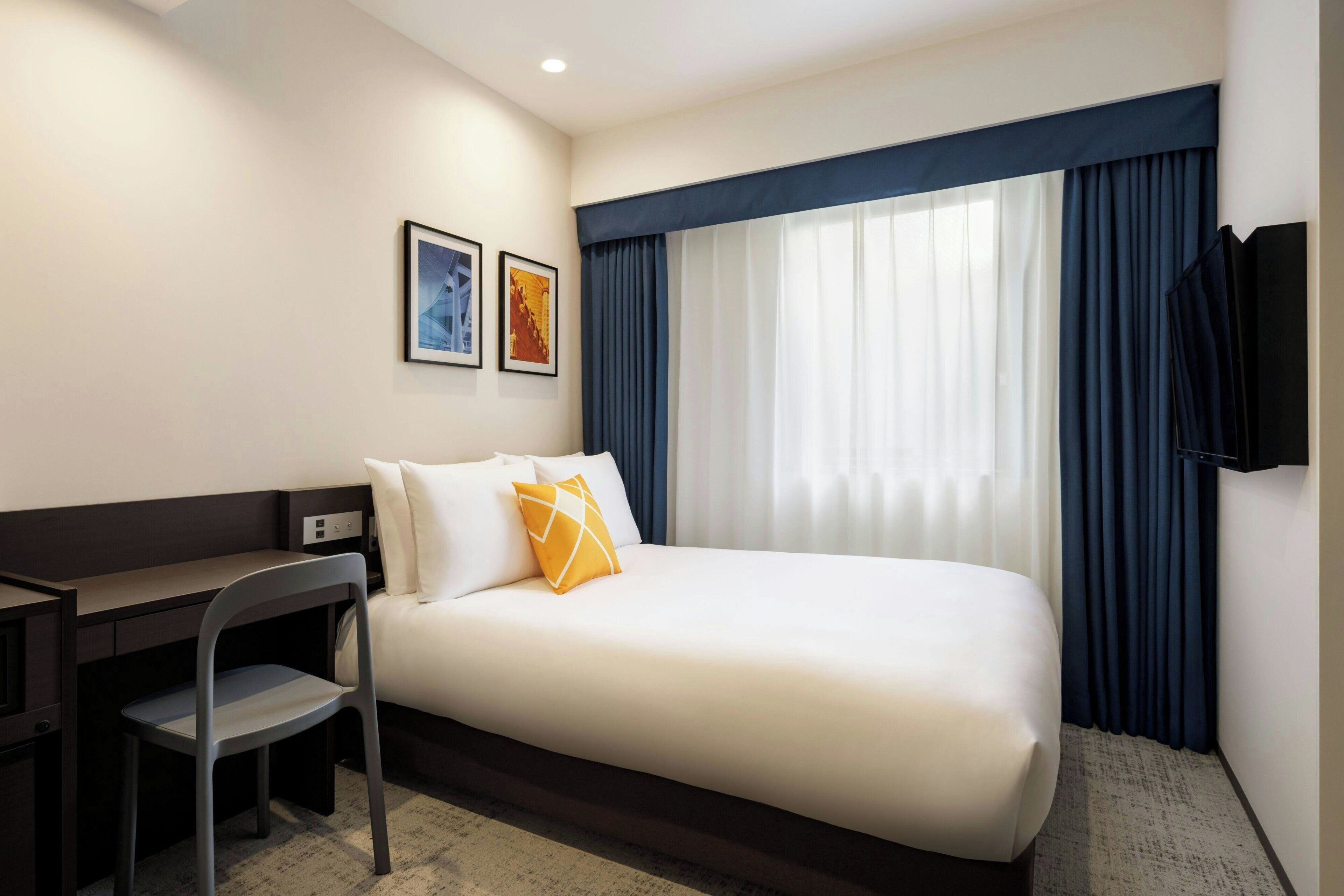 Una habitación de hotel con cama, escritorio, silla, televisor y dos cuadros en la pared.