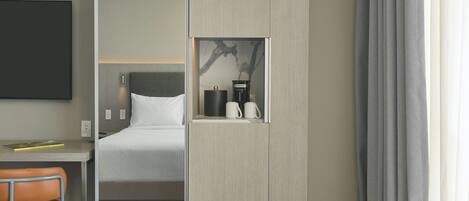Quarto de hotel com cama, secretária, cadeira, máquina de café e televisão presa à parede.