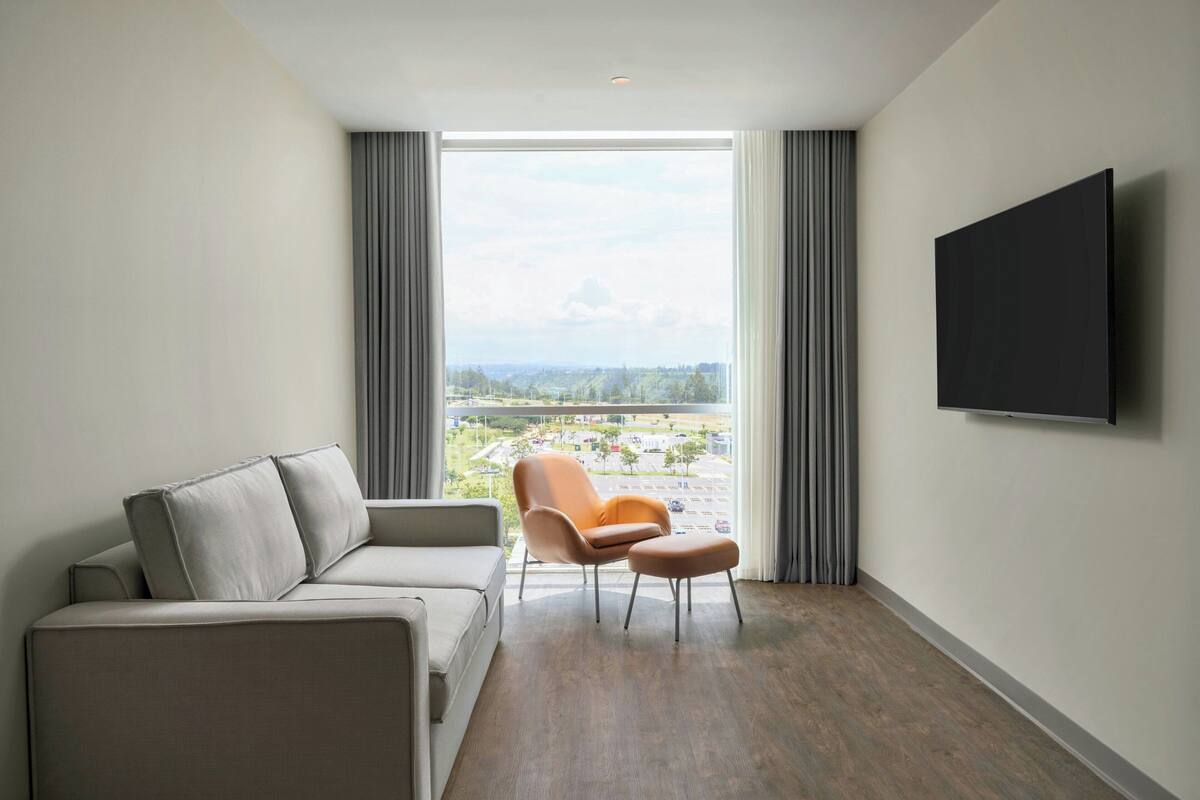 Una habitación de hotel moderna con sofá, sillón, reposapiés y un amplio ventanal con vistas a la ciudad.