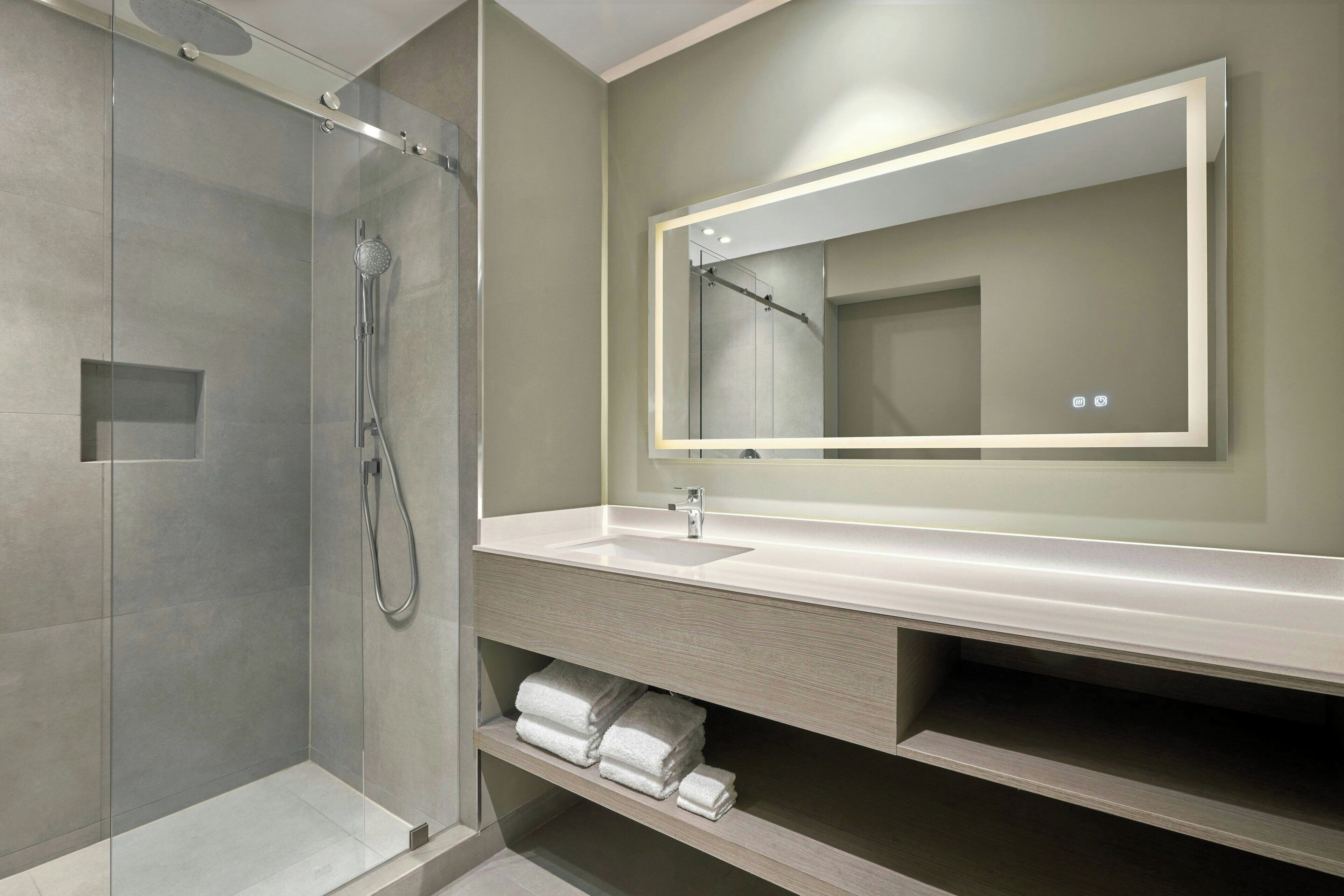 Un baño moderno con ducha de vidrio, un espejo grande y tocador con lavamanos y espacio para toallas.