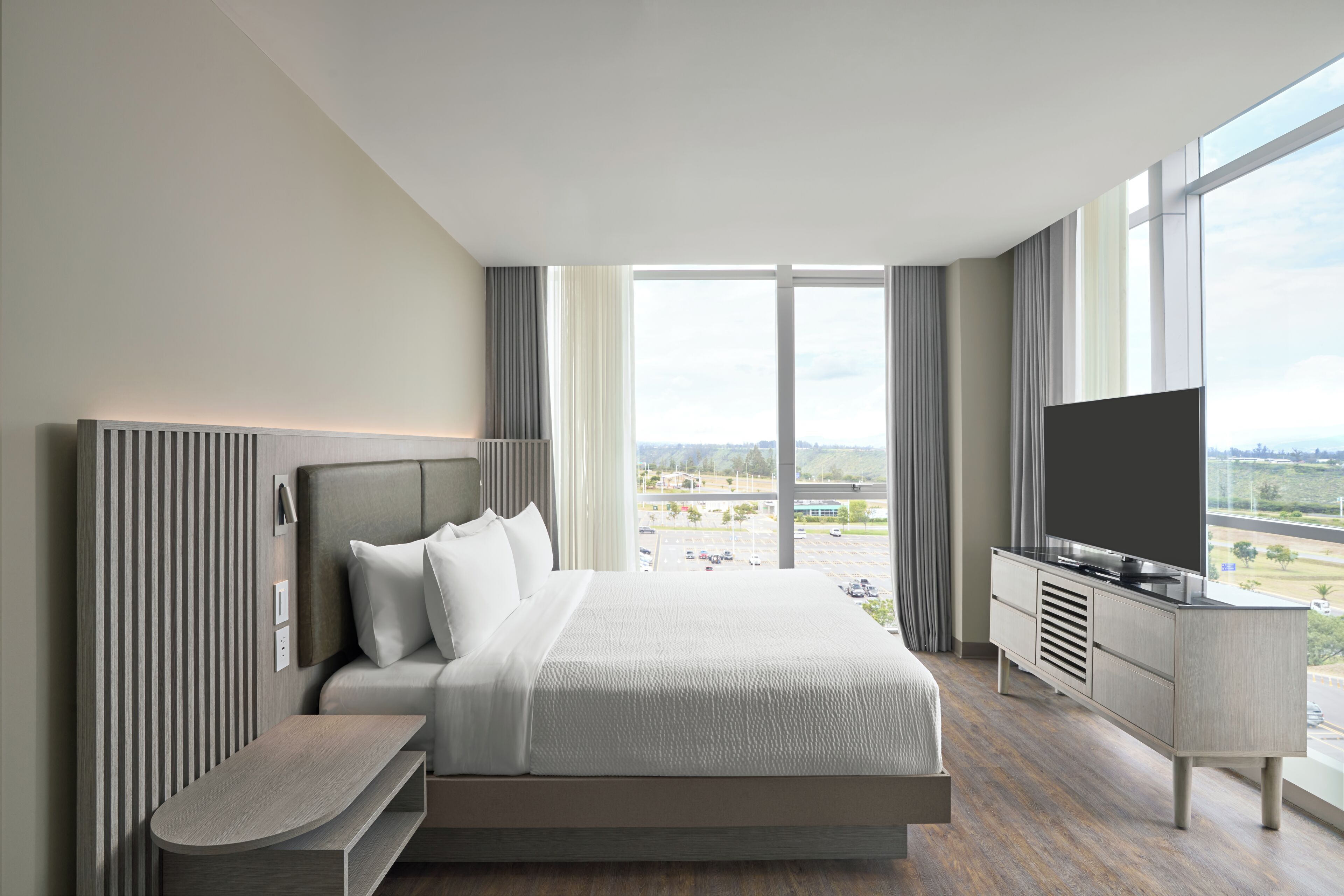 A modern hotel room with a large bed, a flat-screen TV, a desk, and a view of a cityscape through large windows.