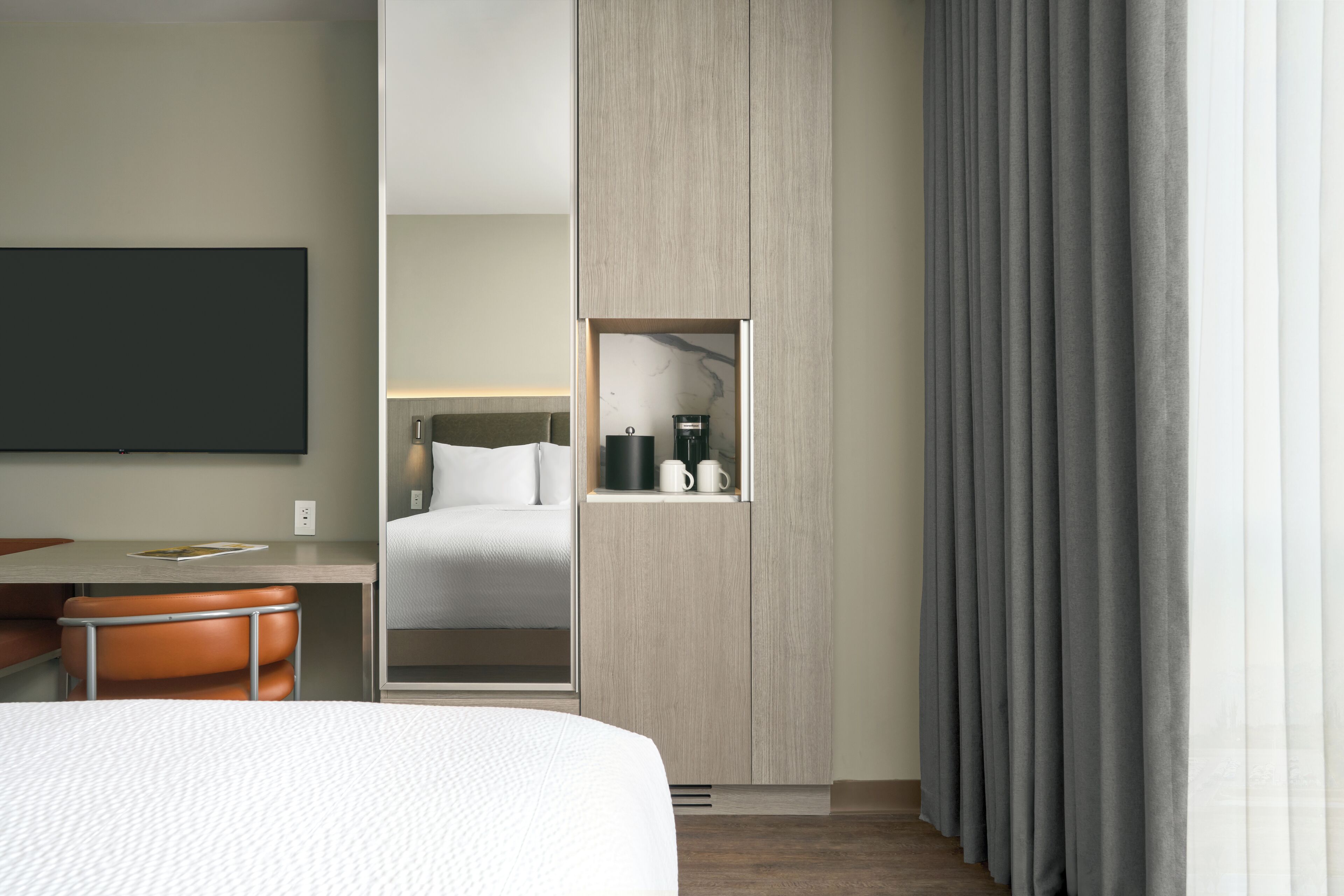 Habitació d'hotel moderna amb un llit, una taula de treball i una cadira. Hi ha un armari, un mirall i una finestra amb cortines.