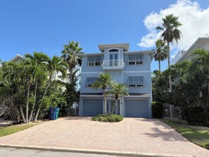 A two-story residential building with a central garage, surrounded by palm trees and a brick driveway.