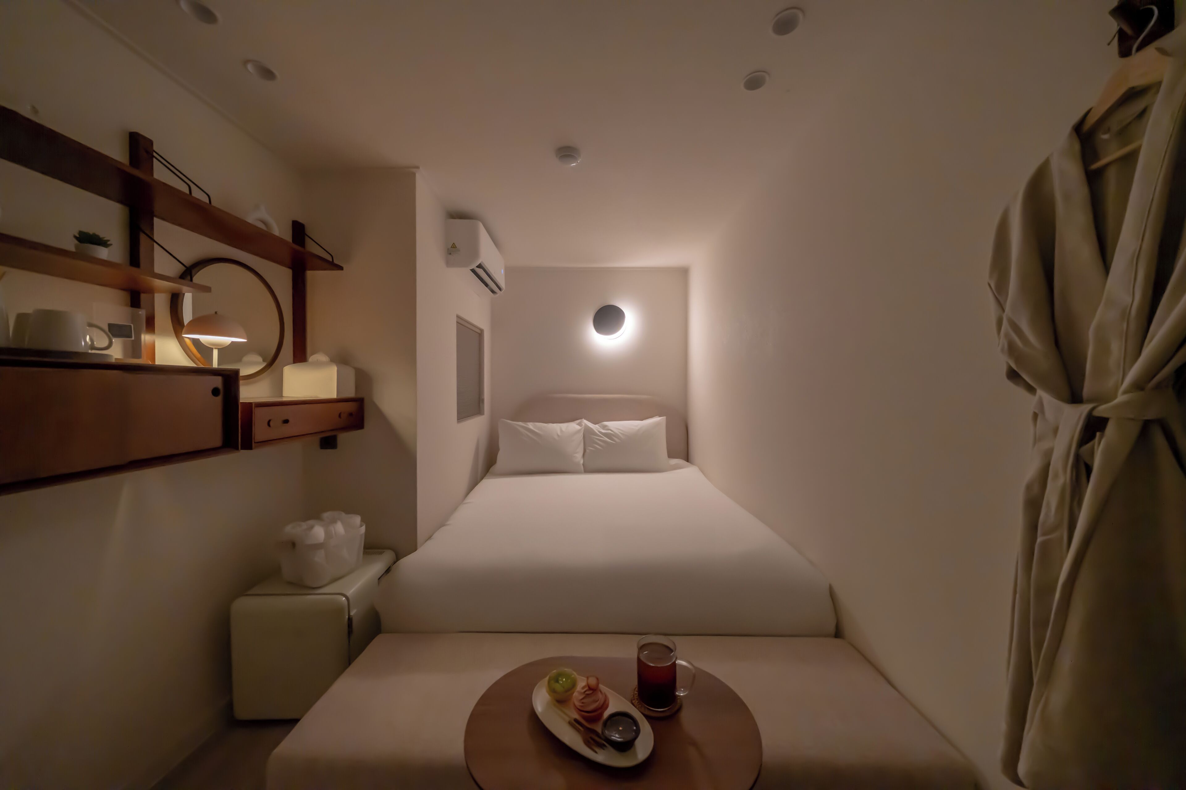 A compact hotel room with a bed, bedside table, and a robe hanging on the wall.