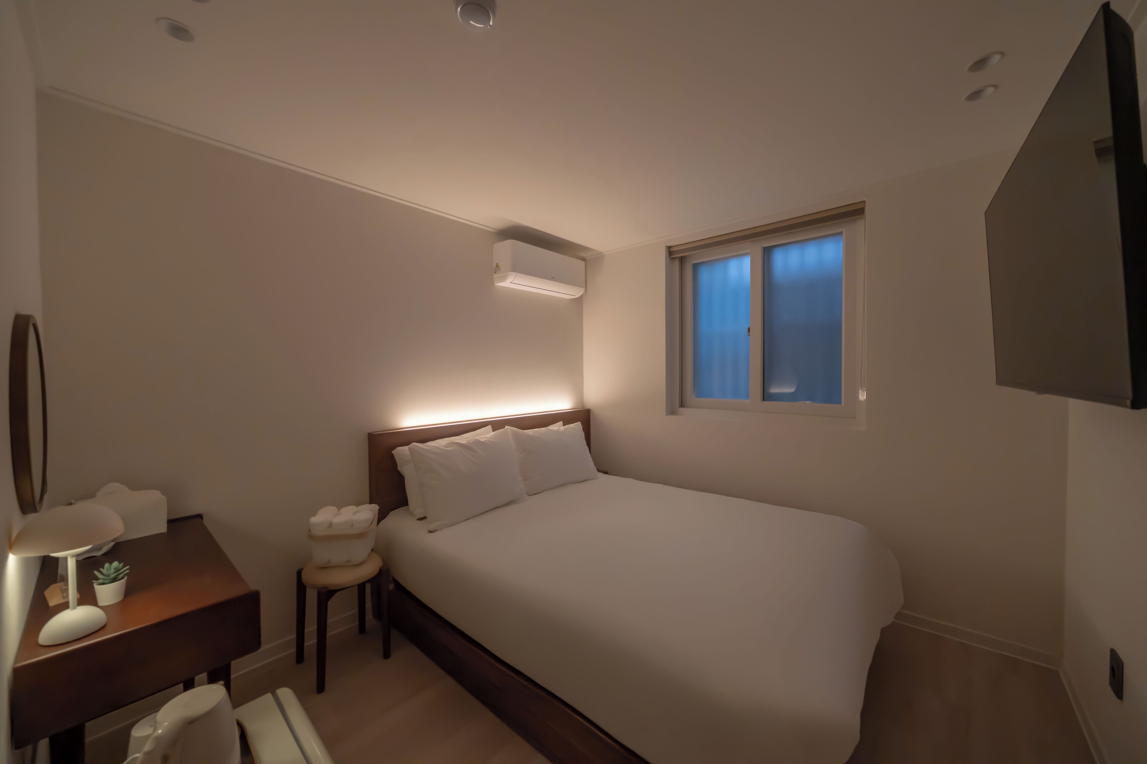 A bedroom with a large bed, a bedside table with a lamp, a small stool, and a window with blinds.