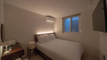 A bedroom with a large bed, a bedside table with a lamp, a small stool, and a window with blinds.