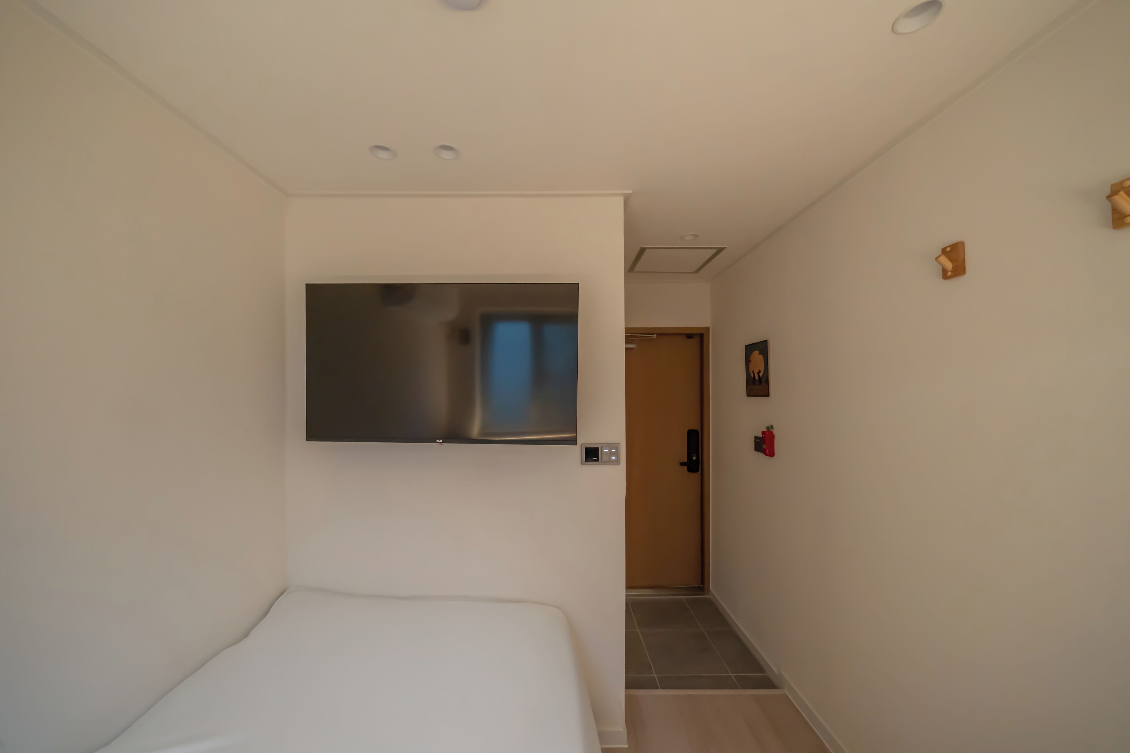 Una habitación de hotel con una cama, un televisor montado en la pared, una puerta que da a un pasillo y un estante pequeño fijado a la pared.