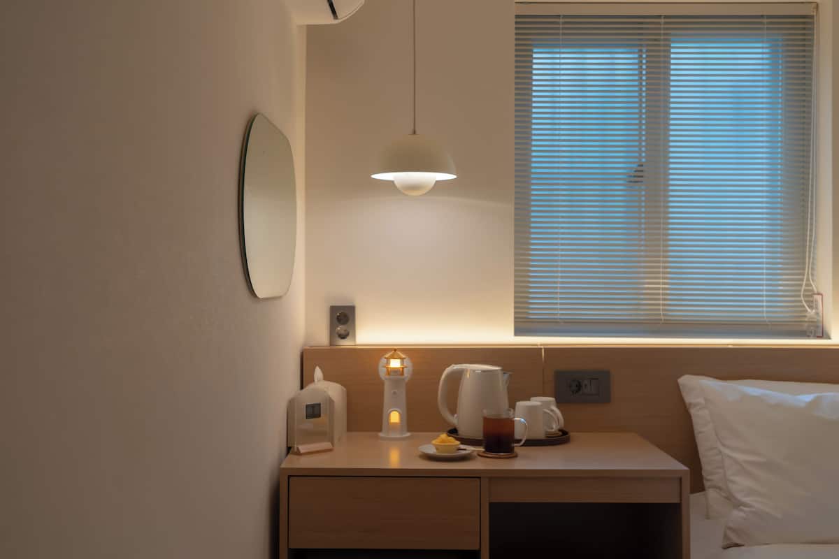 Une chambre d’hôtel équipée d’une table de chevet, d’une lampe, d’une bouilloire, d’une tasse et d’une fenêtre avec des stores.