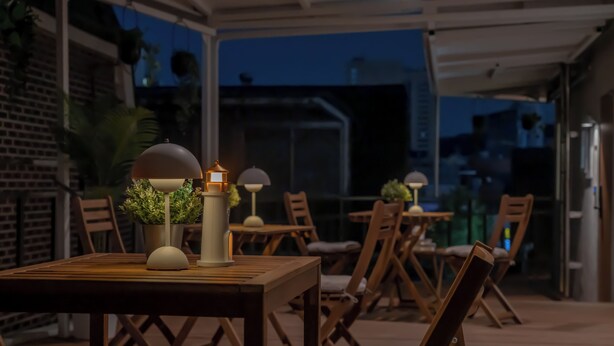 Een terras buiten met houten tafels en stoelen, plantenbakken en decoratieve lampen.