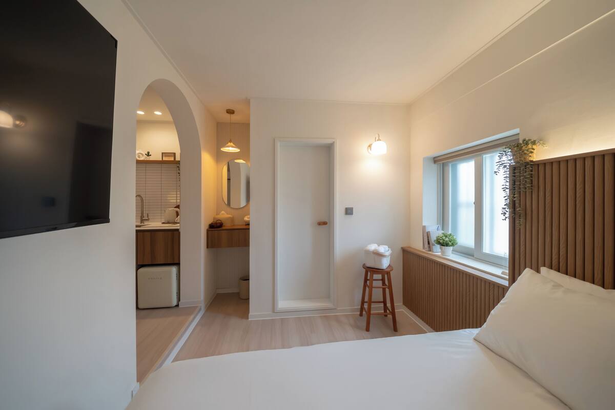Une chambre d’hôtel de taille réduite, comprenant un lit, un petit tabouret, une fenêtre avec des stores, un radiateur et, au fond, une kitchenette.
