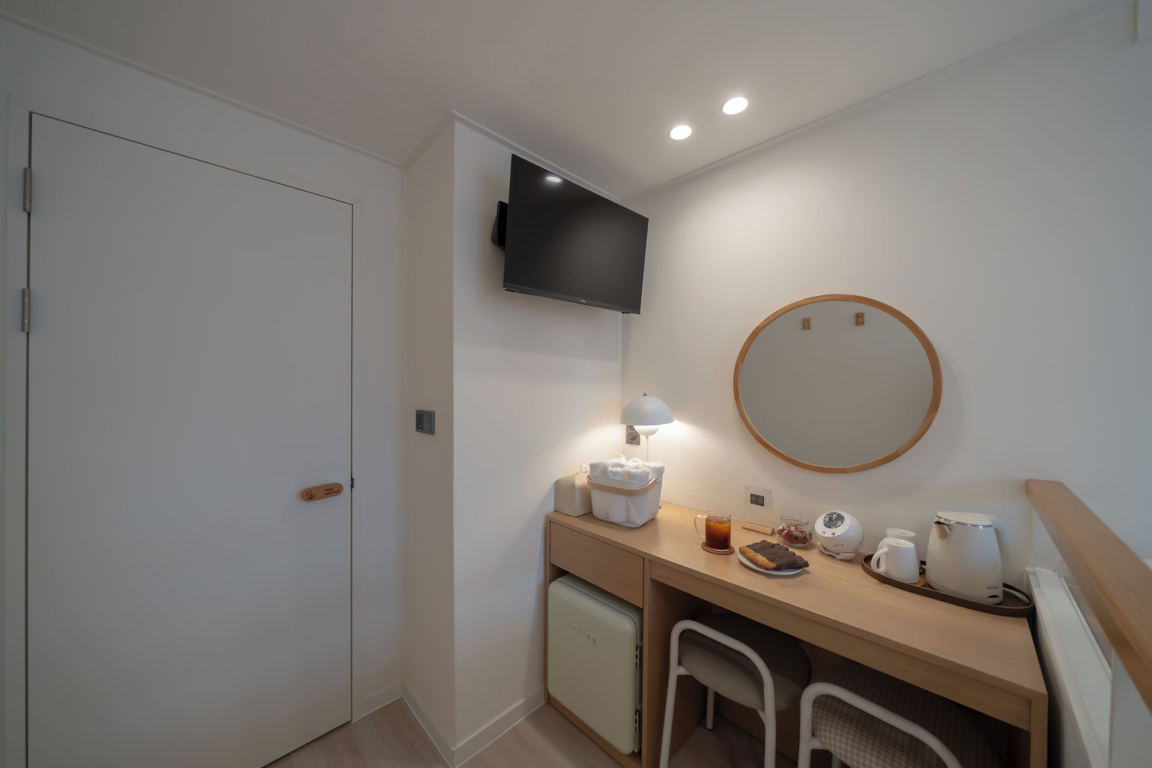 A compact hotel room with a bed, a desk with a lamp, a small refrigerator, a mirror, and a television mounted on the wall.