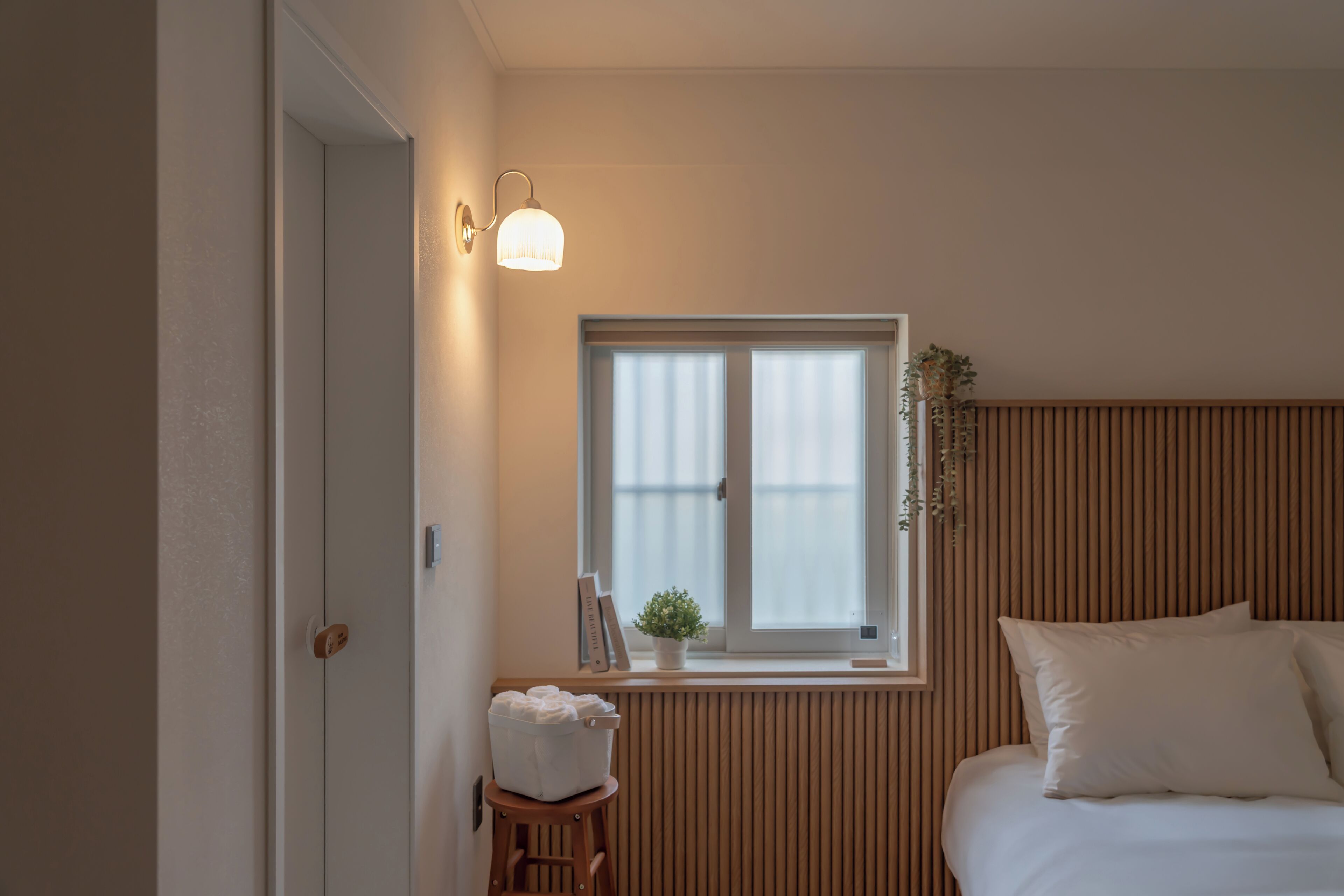 A bedroom with a bed, a window with blinds, a radiator, a small stool, and a wall-mounted light fixture.
