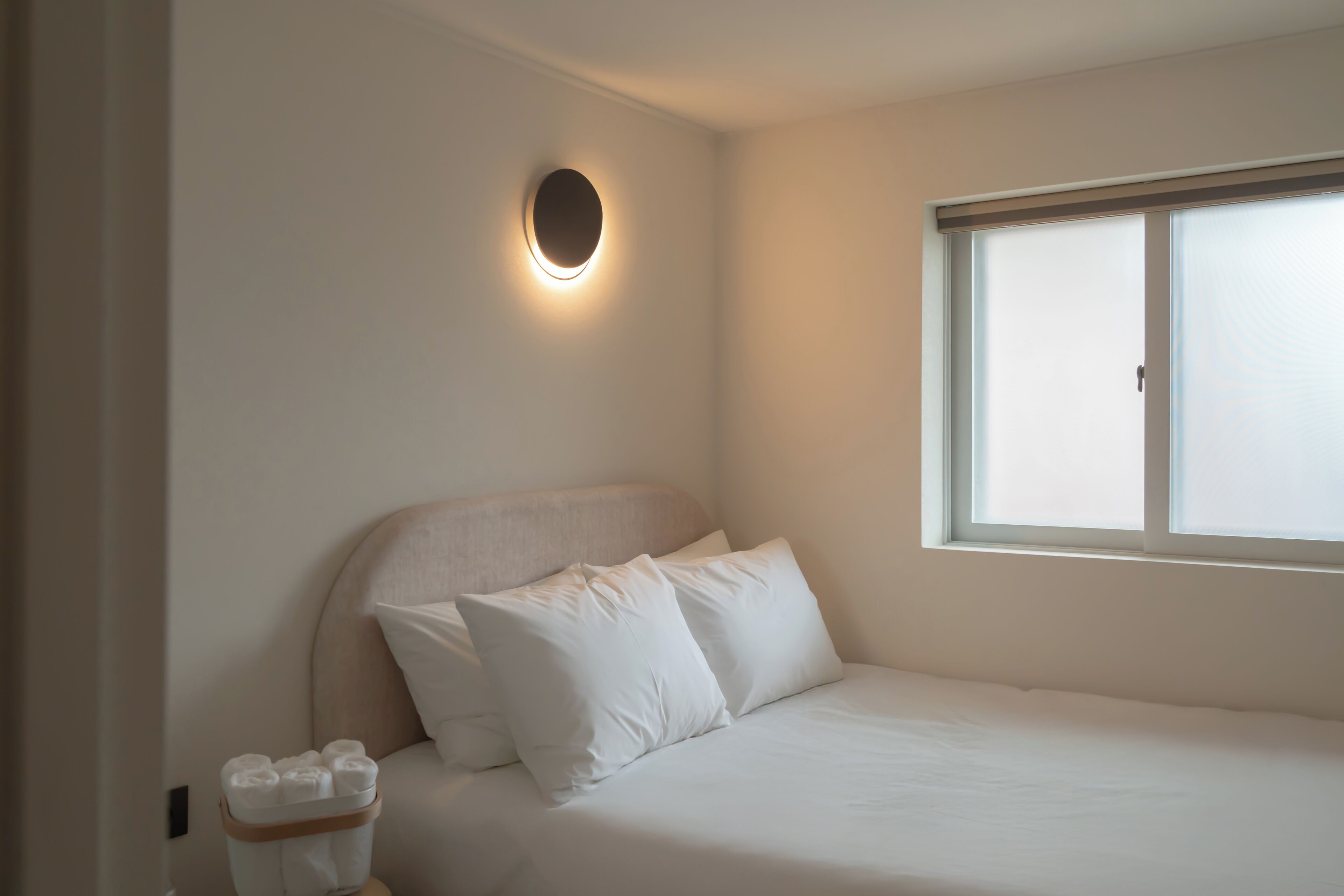 A bedroom with a bed, pillows, a bedside table with toiletries, a wall-mounted light, and a window.