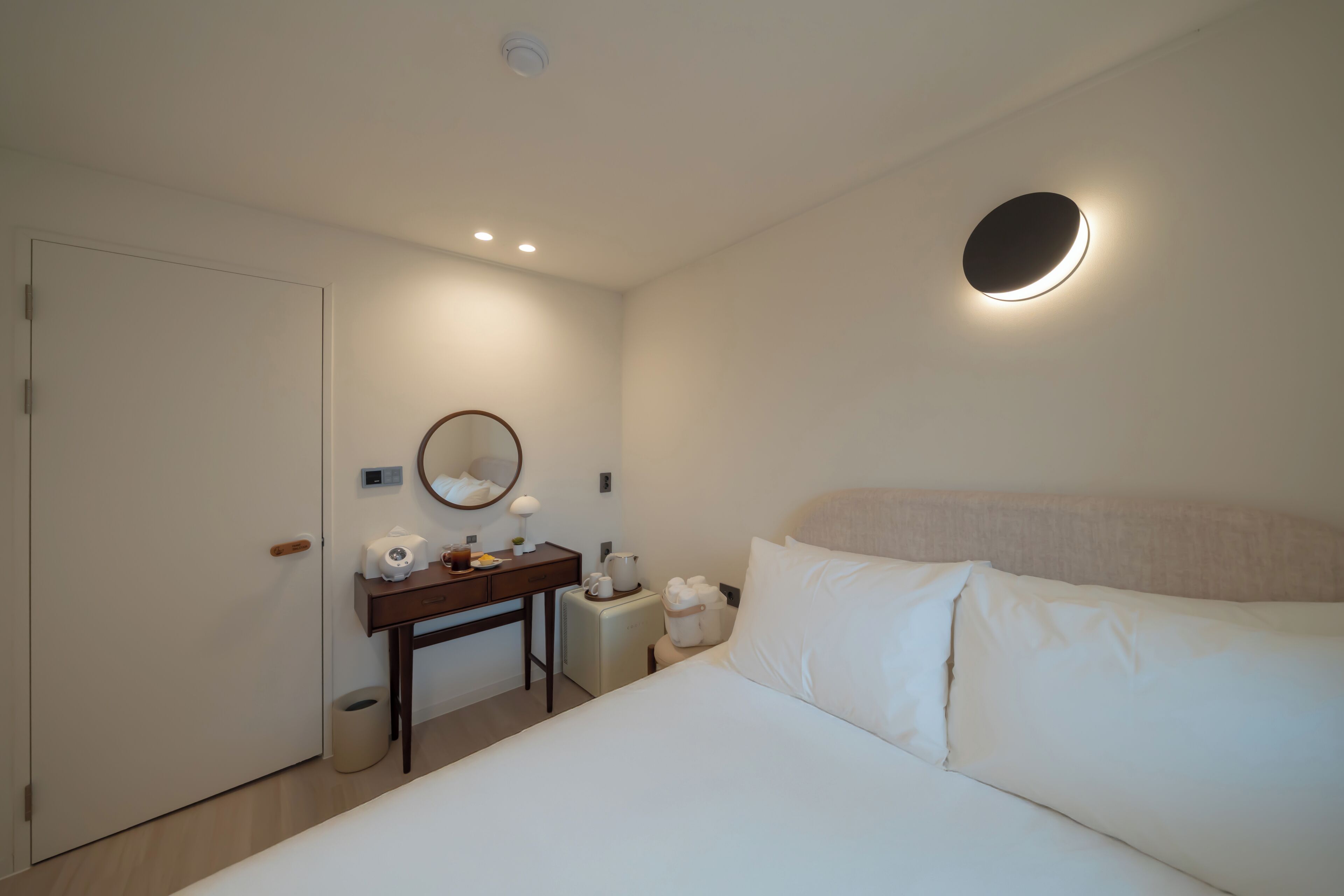 Habitación de hotel con cama, escritorio con espejo, una mesita con tetera y un cubo de basura.