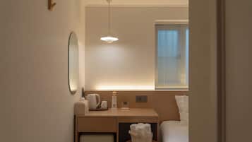 Habitación de hotel pequeña y moderna con cama, mesita de noche y espejo. Cuenta con ventana con persianas, una lámpara en el techo y un pequeño calentador.