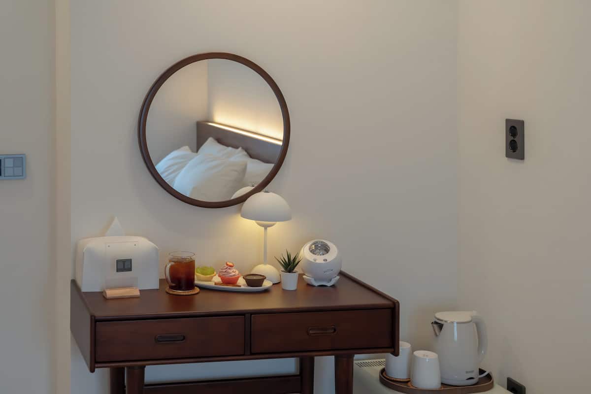 Une chambre d’hôtel avec un bureau en bois, un miroir, une lampe, une machine à café, une théière et une petite plante.