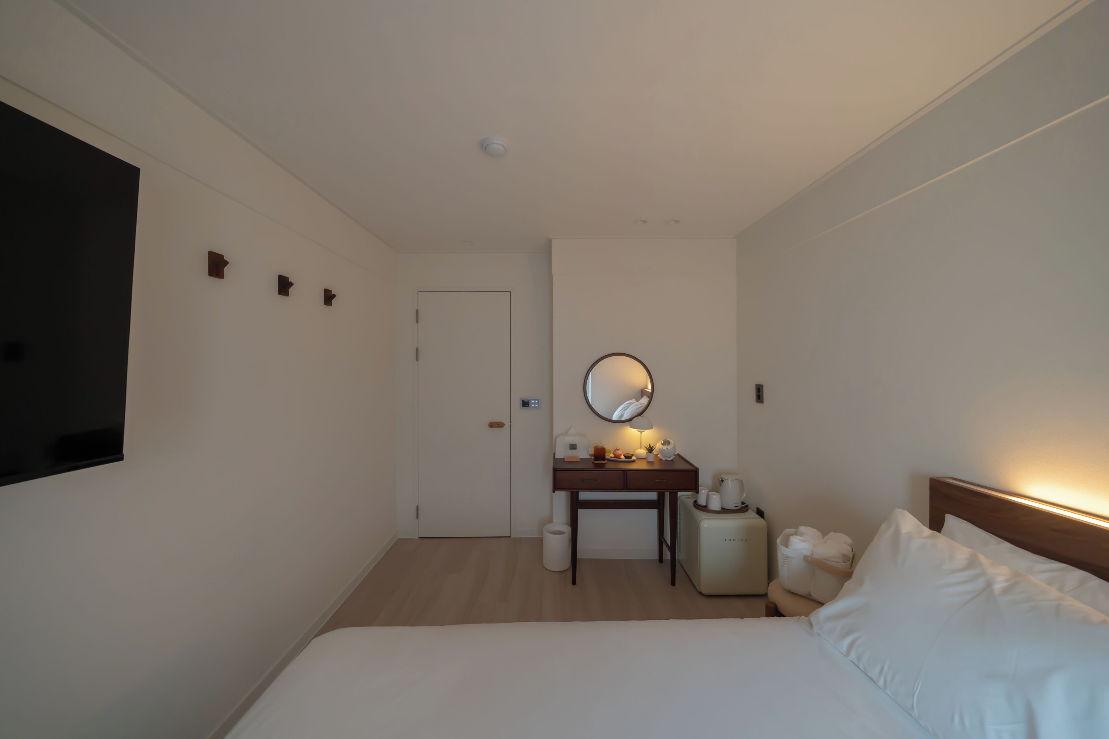 A hotel room with a bed, a desk with a mirror, a small table with a lamp, and a TV mounted on the wall.