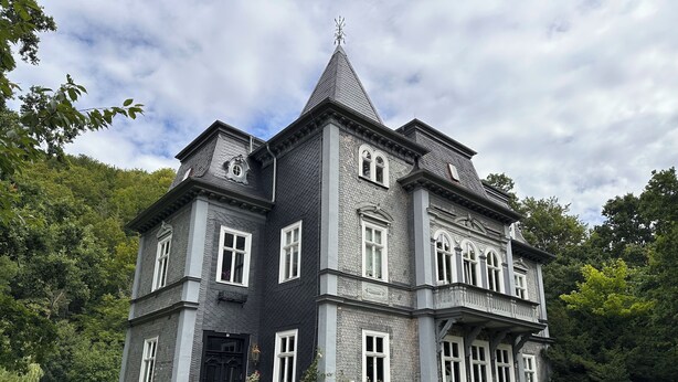 Ein großes, dunkelgraues Mehrgeschosshaus mit weißen Verzierungen und zahlreichen Fenstern, umgeben von üppigem Grün.