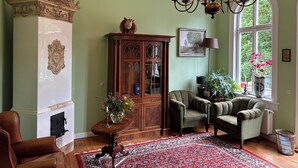 Ein Zimmer mit Kamin, einem Holzschrank, zwei Sesseln, einer Chandelier und einem großen Fenster mit Blick ins Grüne.